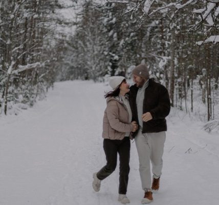 A joyful couple strolls through a snowy forest, embracing winter's beauty and tranquility.