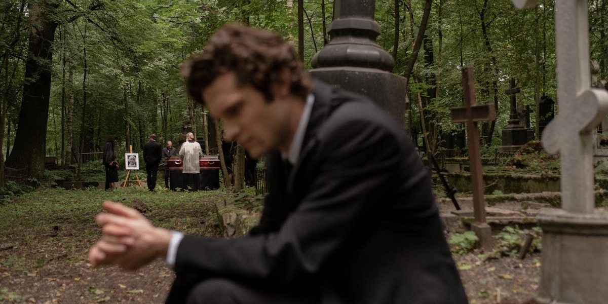 A man in mourning at a funeral surrounded by gravestones in a serene forest cemetery.