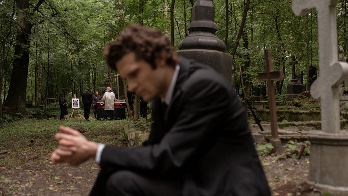 A man in mourning at a funeral surrounded by gravestones in a serene forest cemetery.